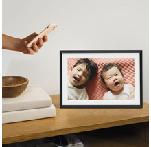 Digitaler Bilderrahmen auf einem Holzschrank mit einem Foto von zwei Kindern, die auf einem rosa Laken liegen. Eine Hand hält ein Smartphone darüber.