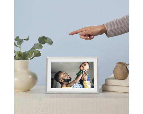 Digitaler Bilderrahmen mit Foto eines Vaters und seines Sohnes auf einem Tisch, daneben eine Vase mit Eukalyptus und eine Hand, die auf den Rahmen zeigt.