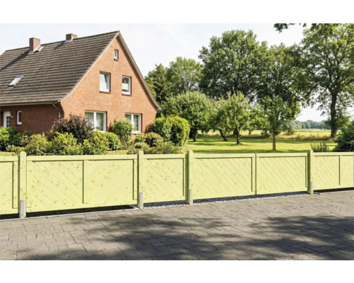 Hellgrüner Gartenzaun aus Holz mit diagonal angeordneten Lamellen vor einem Backsteinhaus mit weitläufigem Garten.