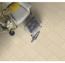 Beige rechteckige Bodenfliesen in einem hellen Raum mit hellgrauem Sessel, Hocker mit grauer Decke und Vase mit Trockenpflanzen.