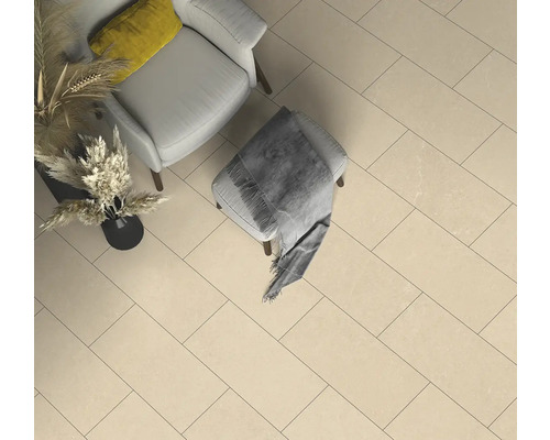 Beige rechteckige Bodenfliesen in einem hellen Raum mit hellgrauem Sessel, Hocker mit grauer Decke und Vase mit Trockenpflanzen.