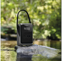 Tragbare Powerstation mit Tragegriff und digitalem Display auf einem Stein in einem Bach vor einem unscharfen Waldhintergrund.