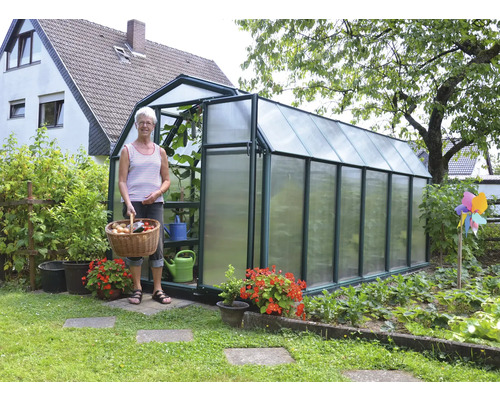 Großes Gewächshaus im Garten mit Hohlkammerplatten aus Polycarbonat und Metallgestell, Frau mit Erntekorb vor dem Eingang.