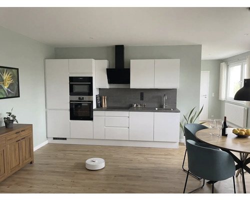 Moderne Wohnküche mit weißen Schränken, grauem Spritzschutz, rundem Holztisch, blauen Stühlen und Saugroboter auf hellem Holzboden.