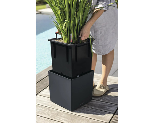 Person hebt einen schwarzen Pflanzeinsatz mit Griffen aus einem quadratischen Pflanzkübel auf einer Holzterrasse.