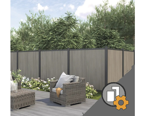 Sichtschutzzaun aus WPC in einem Garten mit Holzterrasse, grauen Lounge-Möbeln aus Geflecht und blühenden Pflanzen vor Bäumen.