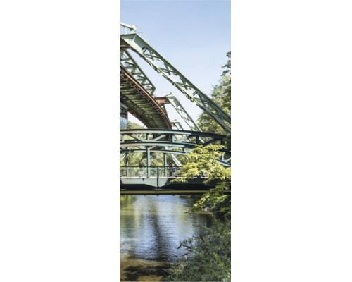 Industrielle Stahlkonstruktion der Wuppertaler Schwebebahn über einem Fluss mit Uferbepflanzung und Fußgängersteg.