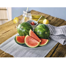 Zwei ganze Wassermelonen und mehrere Wassermelonenscheiben auf einem Teller auf einem Holztisch mit frischem Obst und Getränken.