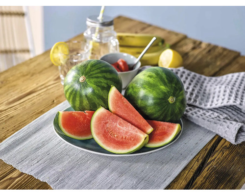 Zwei ganze Wassermelonen und mehrere Wassermelonenscheiben auf einem Teller auf einem Holztisch mit frischem Obst und Getränken.