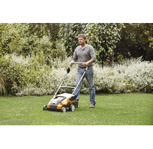 Ein Mann benutzt einen Stihl Vertikutierer auf einer Rasenfläche in einem Garten mit Sträuchern.