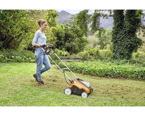 Frau bedient einen Vertikutierer auf einer Rasenfläche in einem Garten.