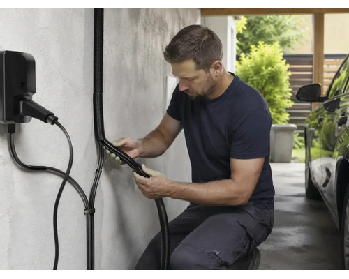 Ein Mann installiert einen schwarzen Wellschlauch als Kabelschutz für eine Wallbox Ladestation an einer Wand in einem Carport.