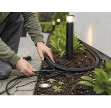 Installation von Gartenbeleuchtung mit schwarzem Wellrohr und Außenkabeln in einem Beet neben Pollerleuchten.