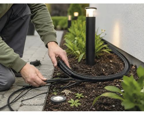 Installation von Gartenbeleuchtung mit schwarzem Wellrohr und Außenkabeln in einem Beet neben Pollerleuchten.