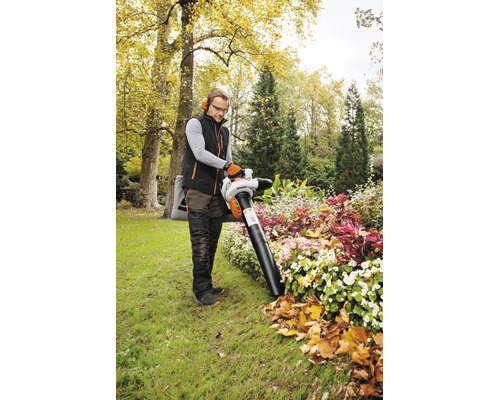 Ein Mann arbeitet mit einem STIHL Saughäcksler in einem hellen Garten. Er saugt Herbstlaub von einem bepflanzten Blumenbeet. Er trägt Schutzausrüstung.