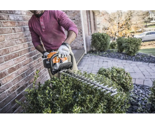 Person schneidet einen Busch mit einer Stihl Benzin-Heckenschere neben einer Ziegelwand im Garten.