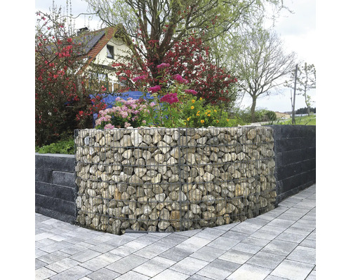 Geschwungene Gabione gefüllt mit Natursteinen als Gartenmauer, umgeben von blühenden Pflanzen und einem gepflasterten Weg.