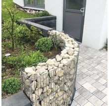Gebogene Gabione als Beeteinfassung im Garten, gefüllt mit hellen Natursteinen neben einem gepflasterten Weg.