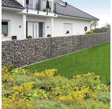 Lange Gabionenwand aus Natursteinen in einem hellen Garten mit Rasenfläche vor einem Wohnhaus mit Balkon.