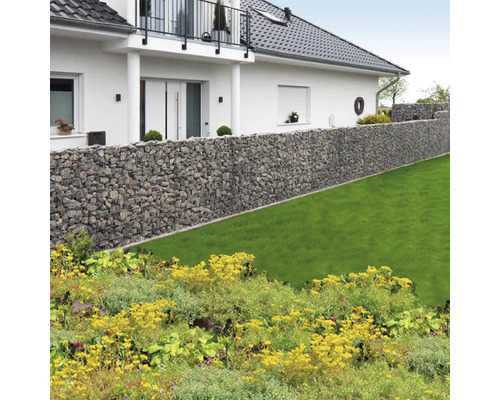 Lange Gabionenwand aus Natursteinen in einem hellen Garten mit Rasenfläche vor einem Wohnhaus mit Balkon.