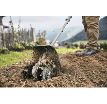 Eine Person benutzt einen Bodenfräsen-Aufsatz, um die Erde in einem Garten mit Zaun und Bergen im Hintergrund aufzulockern.