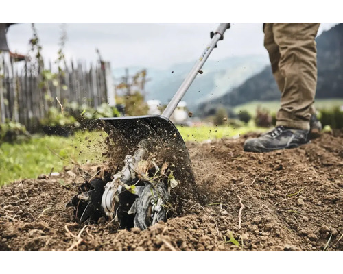 Eine Person benutzt einen Bodenfräsen-Aufsatz, um die Erde in einem Garten mit Zaun und Bergen im Hintergrund aufzulockern.