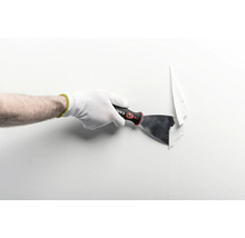 Hand mit Arbeitshandschuh nutzt einen Malerspachtel zum Entfernen von Tapete von einer Wand.