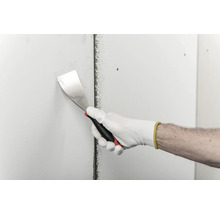 Eine Hand in einem Arbeitshandschuh trägt mit einem Spachtel Spachtelmasse auf eine Trockenbauwand auf.