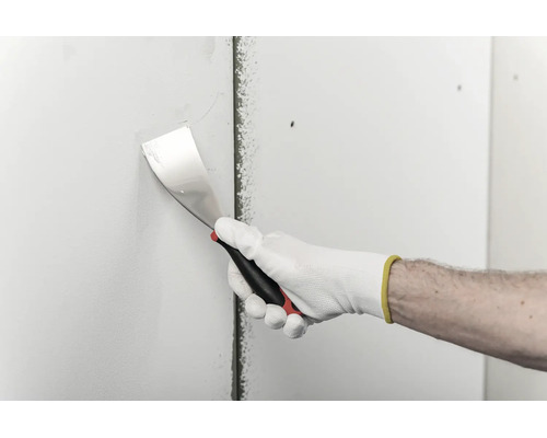 Eine Hand in einem Arbeitshandschuh trägt mit einem Spachtel Spachtelmasse auf eine Trockenbauwand auf.
