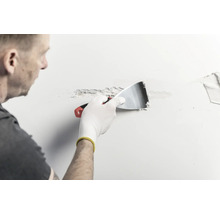 Eine Person trägt mit einem Metallspachtel Spachtelmasse auf eine Wand auf.