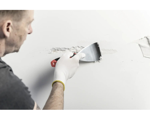 Eine Person trägt mit einem Metallspachtel Spachtelmasse auf eine Wand auf.