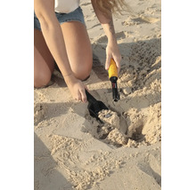 Eine Person nutzt einen Hand-Metalldetektor mit Minelab Logo und eine Sandschaufel, um eine Münze im Sand am Strand auszugraben.