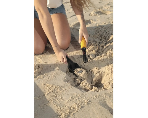 Eine Person nutzt einen Hand-Metalldetektor mit Minelab Logo und eine Sandschaufel, um eine Münze im Sand am Strand auszugraben.