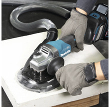 Eine Person in Arbeitshandschuhen bedient einen Akku-Winkelschleifer mit Absaughaube und Saugschlauch auf einer Platte. Makita Logo.