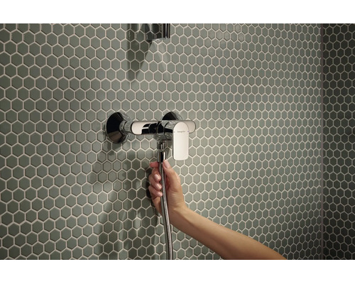 Eine Hand hält einen Duschschlauch an einer Hansgrohe Einhebel Duscharmatur vor einer Wand mit sechseckigen Mosaikfliesen.