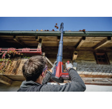 Eine Person reinigt eine Dachrinne mit einem Einhell Akku Dachrinnenreinigungsset an einem rustikalen Holzhaus bei sonnigem Wetter.