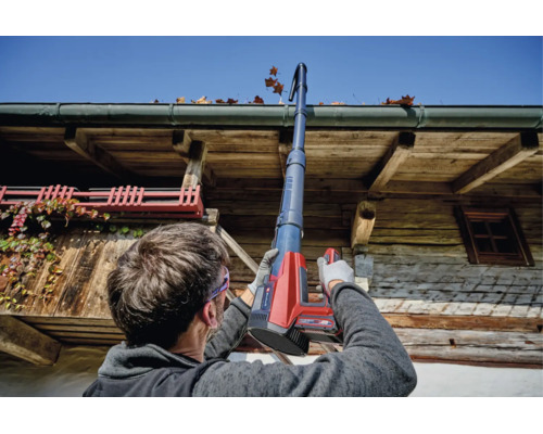 Eine Person reinigt eine Dachrinne mit einem Einhell Akku Dachrinnenreinigungsset an einem rustikalen Holzhaus bei sonnigem Wetter.