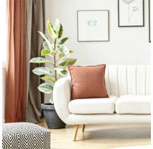 Ein helles Wohnzimmer mit weißem Sofa, einem dekorativen Kissen in Rostbraun, einer Zimmerpflanze im Topf und gemustertem Hocker.