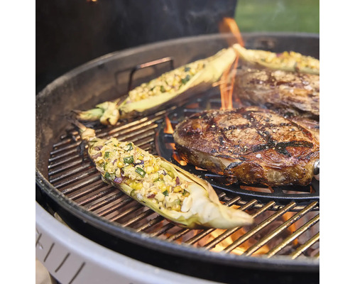 Ein Kugelgrill mit brennender Holzkohle, auf dem ein Rindersteak und gefüllte Maiskolben direkt über dem Feuer gegrillt werden.