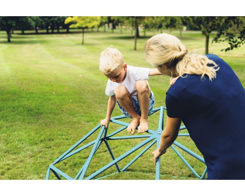 Junge klettert auf Klettergerüst im Garten mit Unterstützung einer erwachsenen Person.