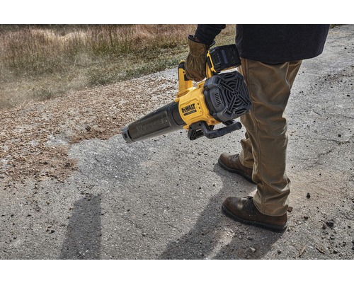 Eine Person bedient einen gelben Akku-Laubbläser von DeWalt auf einer unbefestigten Oberfläche.