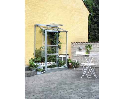 Anlehngewächshaus Halls Altan 2 Blankglas 3 mm 132 x 69 cm Aluminium Anlehn-Gewächshaus aus Glas mit geöffneter Tür, flankiert von Bepflanzung in Töpfen, im Hintergrund eine Gartenmöbelgruppe