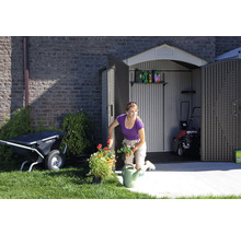 Gartenszene mit einer Frau, die Blumen pflanzt, einem Gartenschuppen mit geöffneten Türen, einer Schubkarre und einem Rasenmäher.