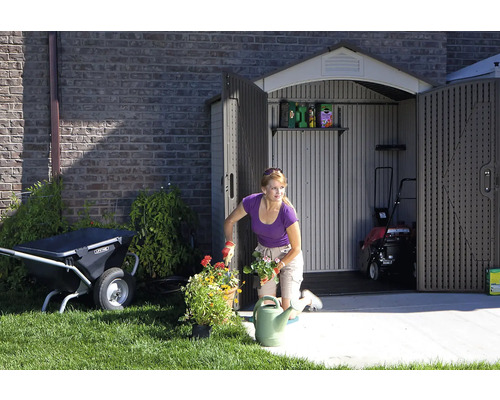 Gartenszene mit einer Frau, die Blumen pflanzt, einem Gartenschuppen mit geöffneten Türen, einer Schubkarre und einem Rasenmäher.