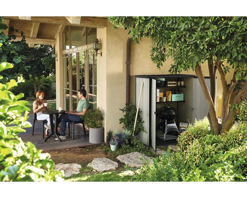 Gartenszene mit einem Gerätehaus, Gartengeräten und einer sitzenden Frau und einem sitzenden Mann