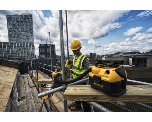 Handwerker mit Schutzhelm und Warnweste arbeitet mit einem Nass-Trockensauger von Dewalt auf einem Baugerüst vor einer Stadtkulisse.