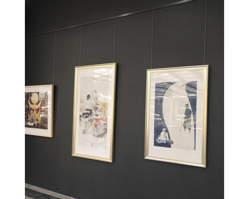 Drei gerahmte Kunstwerke hängen an einer grauen Wand, befestigt mit Seilen