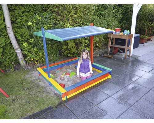 Kind sitzt in buntem Sandkasten mit Sonnendach im Garten.