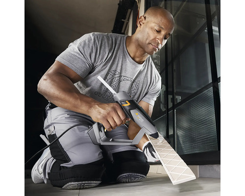 Ein Mann verwendet eine Heißklebepistole zum Anbringen einer Fliese. Die Klebepistole ist grau und schwarz. Die Fliese ist beige.