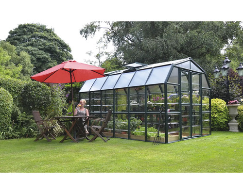 Gewächshaus im Garten mit Pflanzen und Gartengeräten, daneben Tisch und Stühle unter einem Sonnenschirm